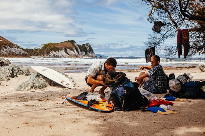 Two men are packing up their beach gear, including a surfboard, beach towels, and a cooler with food and drinks.