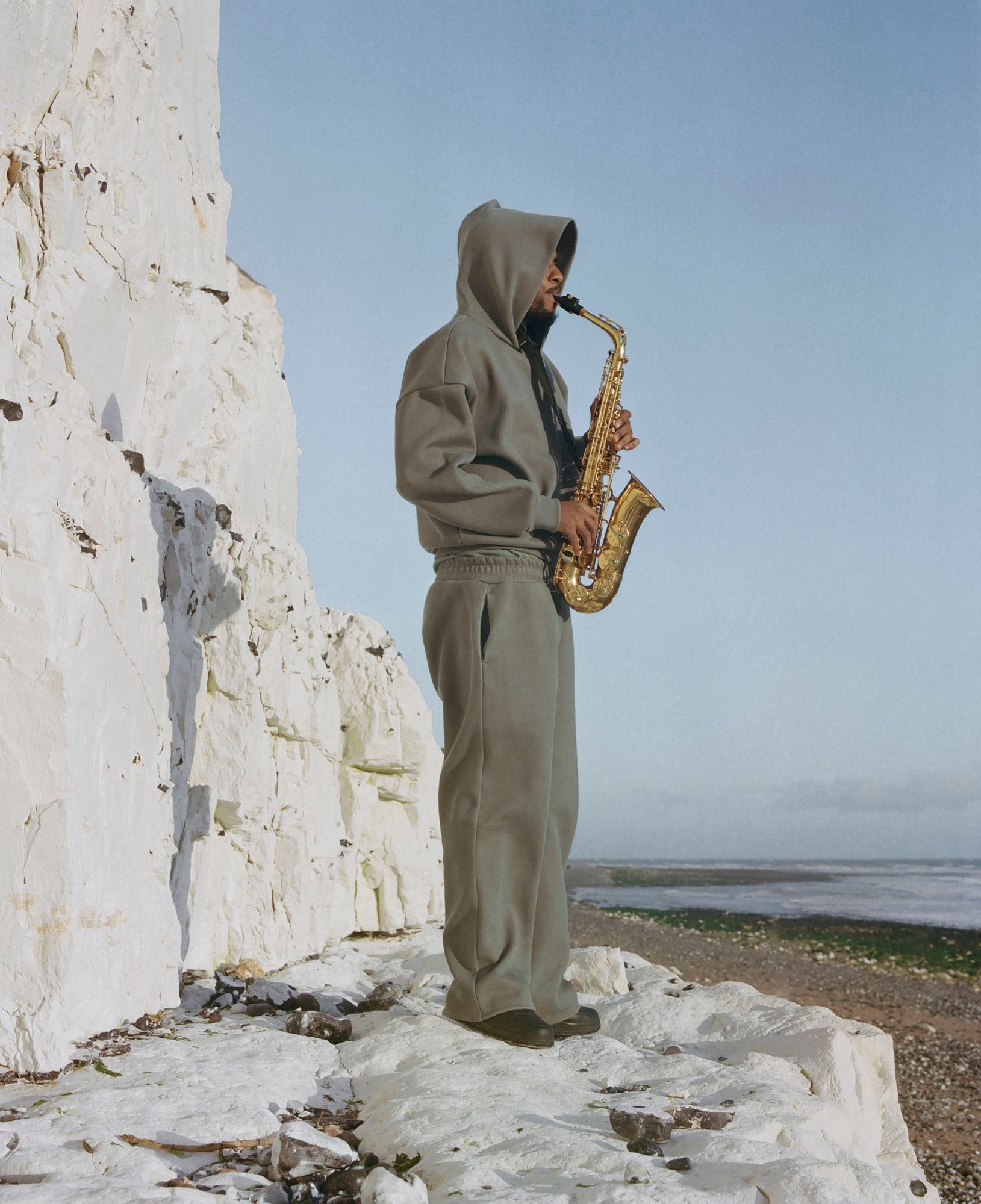 A man in a grey hoodie and sweatpants, playing a saxophone on a rocky beach with a white cliff in the background.
