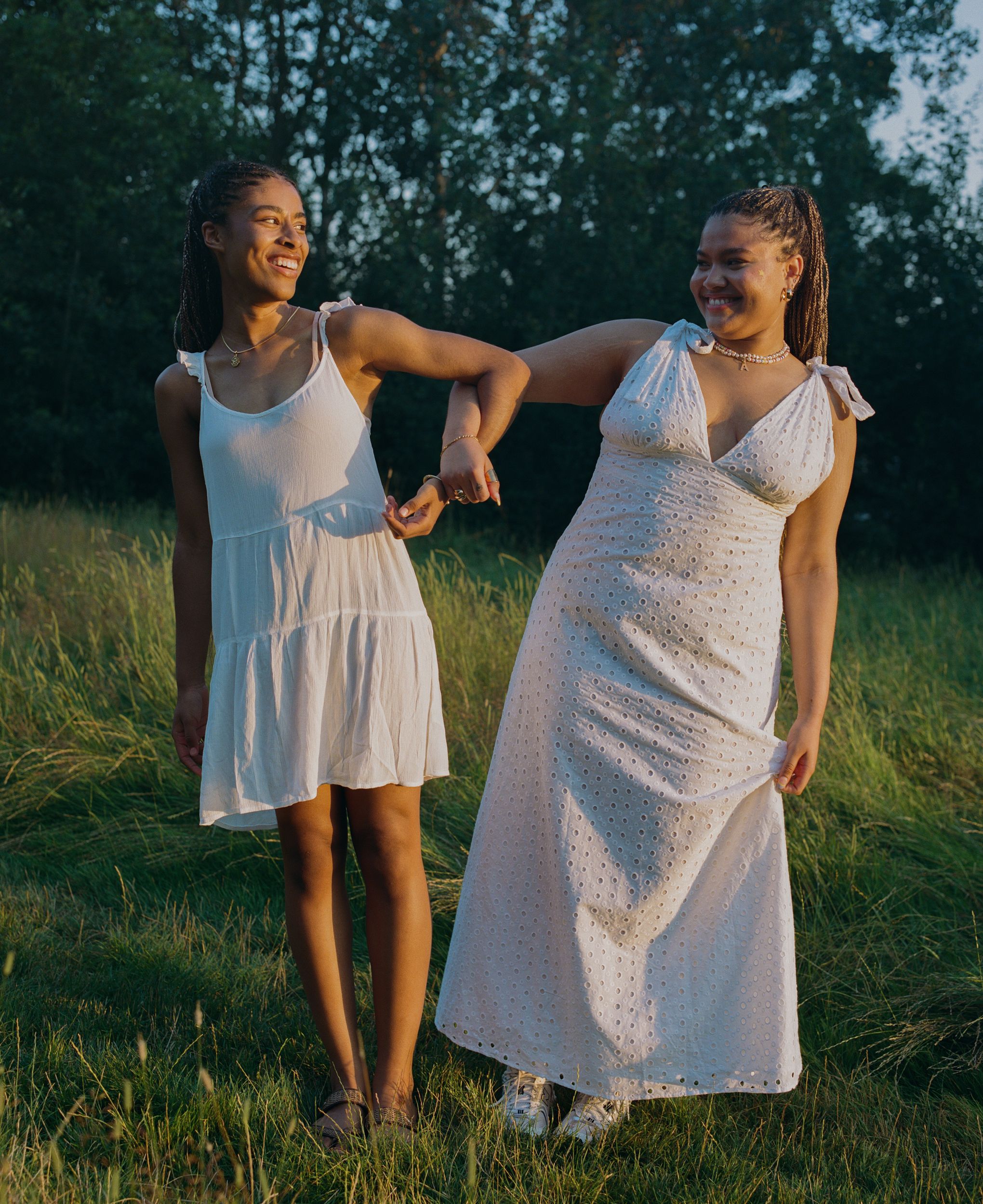 Two young women with braided hair and white sundresses standing in a lush green field, smiling and holding hands.