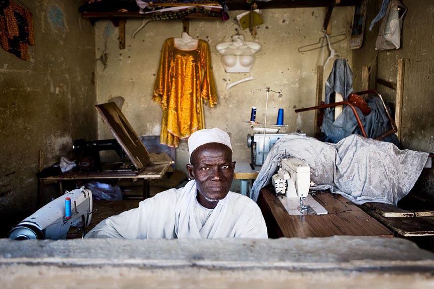 A man in a white robe and turban sits in a small, cluttered sewing room, surrounded by sewing machines, fabric, and various sewing tools.