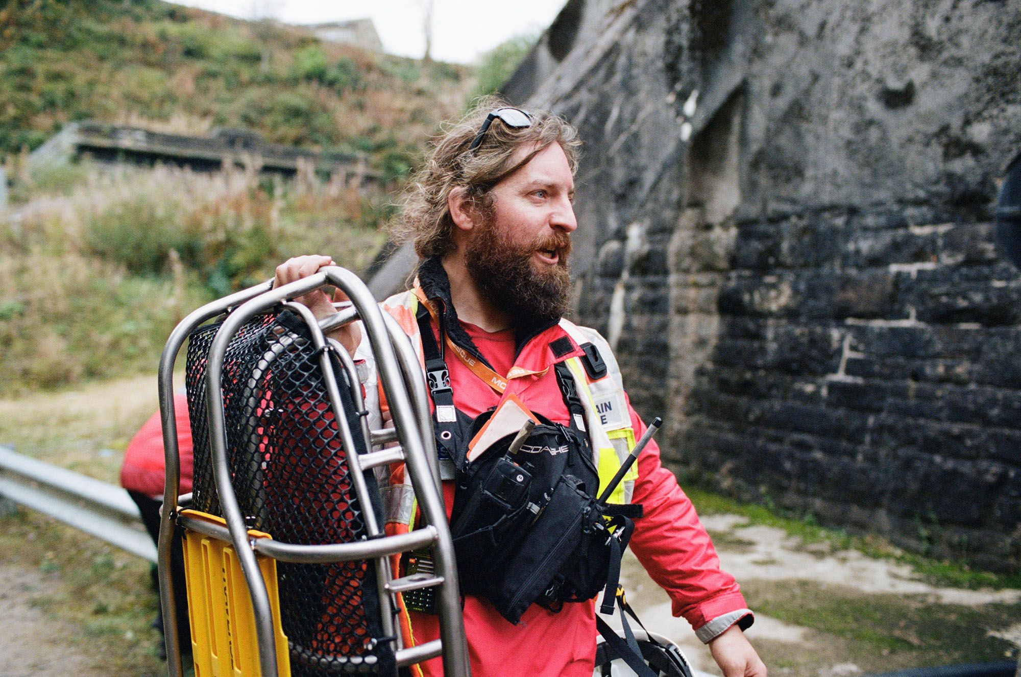 A man with a beard and a red jacket, wearing a safety vest and carrying a rescue basket, standing in front of a stone wall.