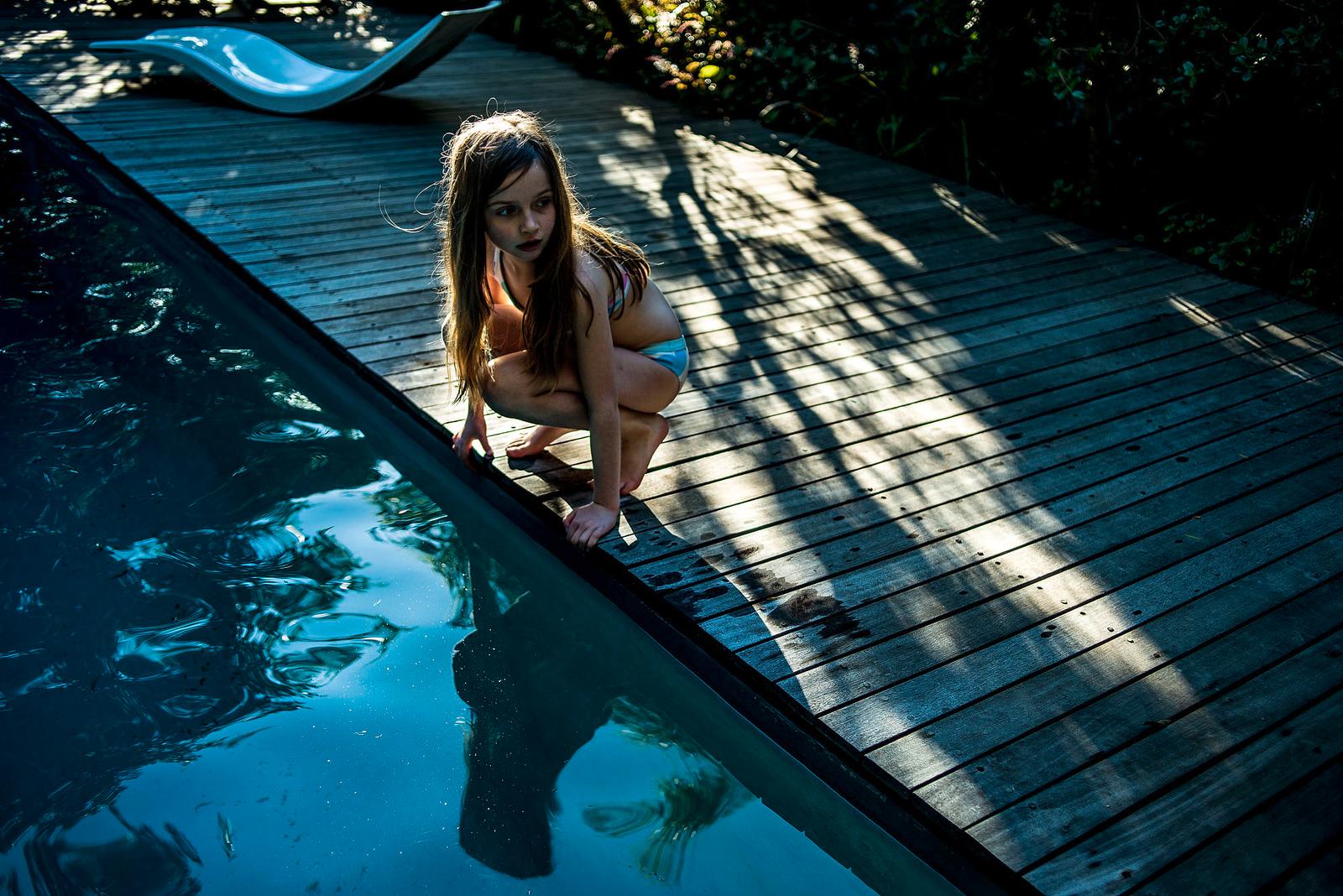 A young girl in a white bikini crouches on the edge of a pool, looking down into the water.