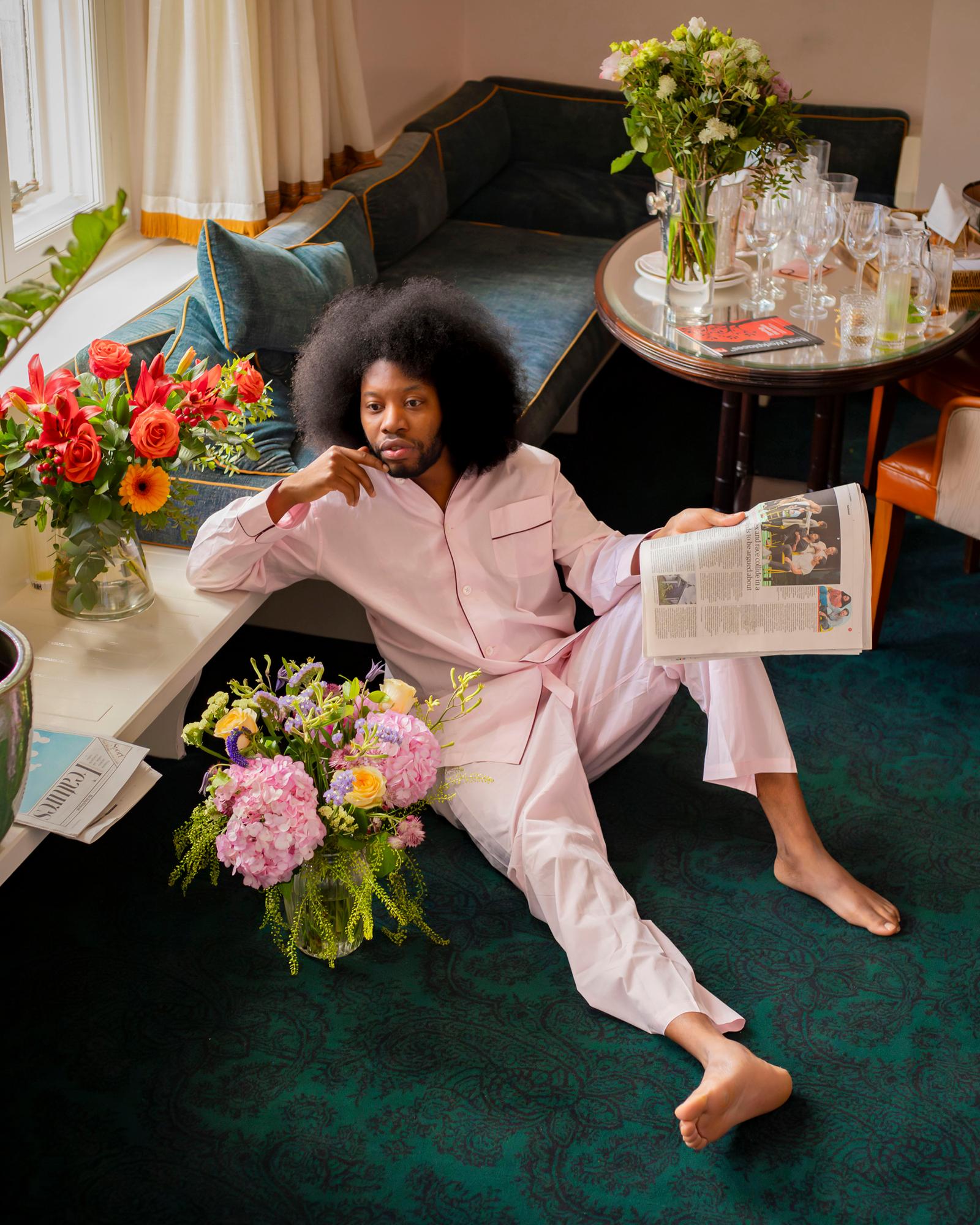 A man with an afro sits on a couch, wearing a pink shirt and white pants, surrounded by flowers and a newspaper.
