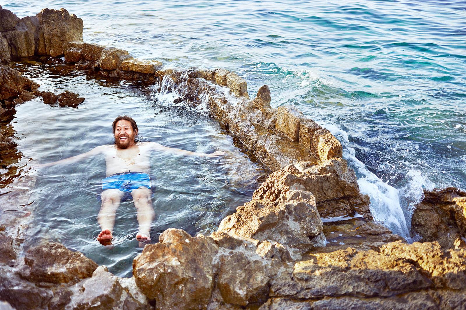 A man in a wetsuit is floating on his back in a rocky, oceanic cove, surrounded by large boulders and small waves.