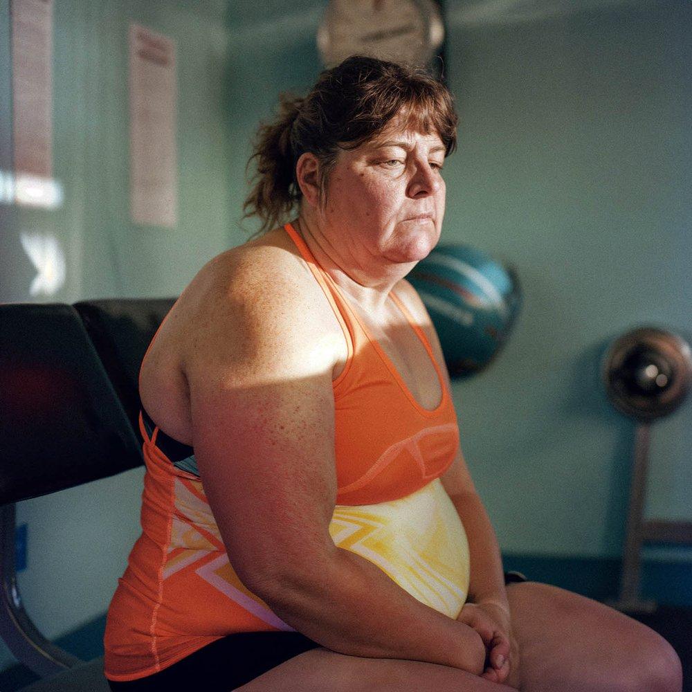 A woman in a bright orange tank top and black shorts sits on a chair, looking unwell and possibly in a gym or fitness center.