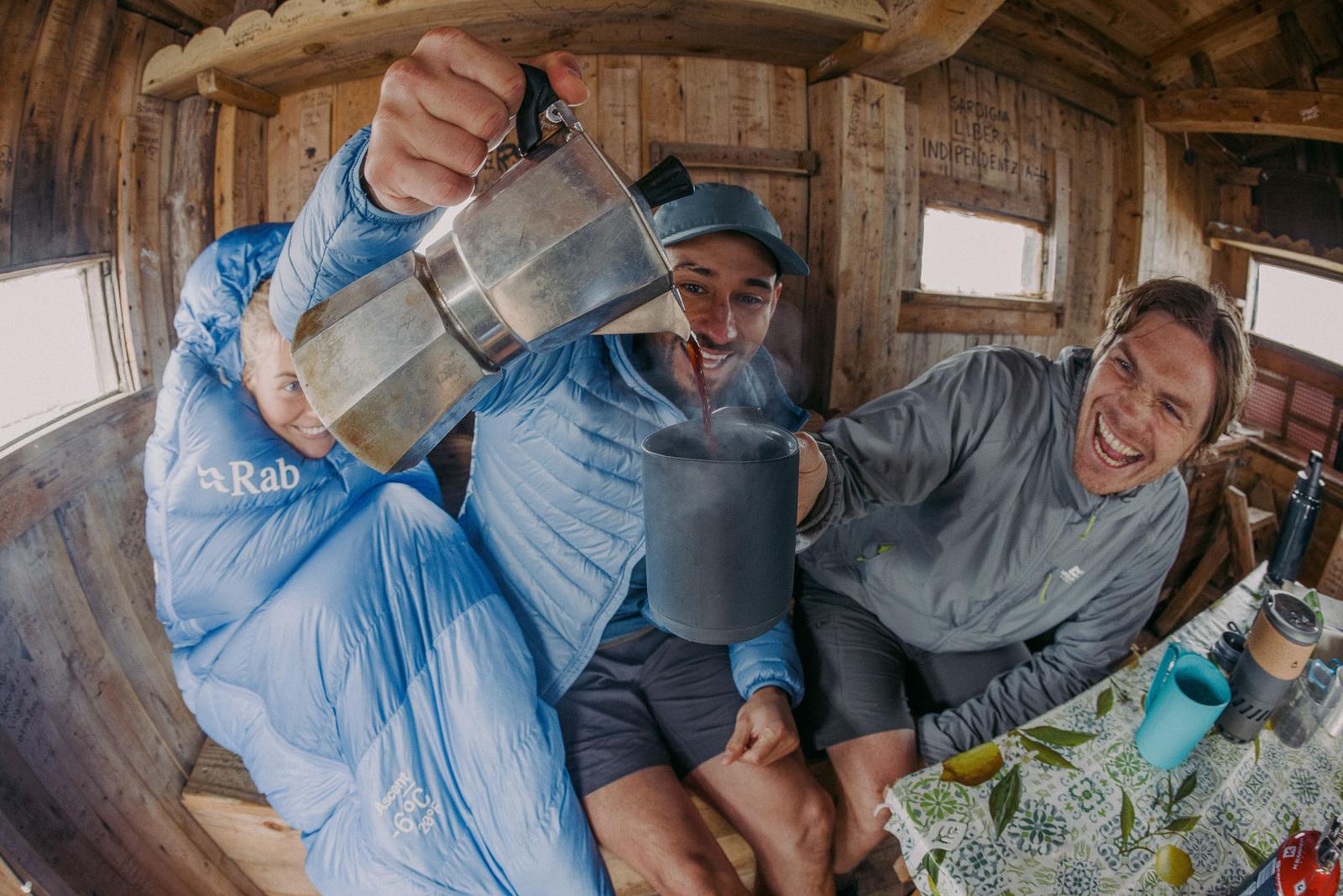 Group of three young people in a mountain hut pouring coffee from a moka pot and laughing james bowden