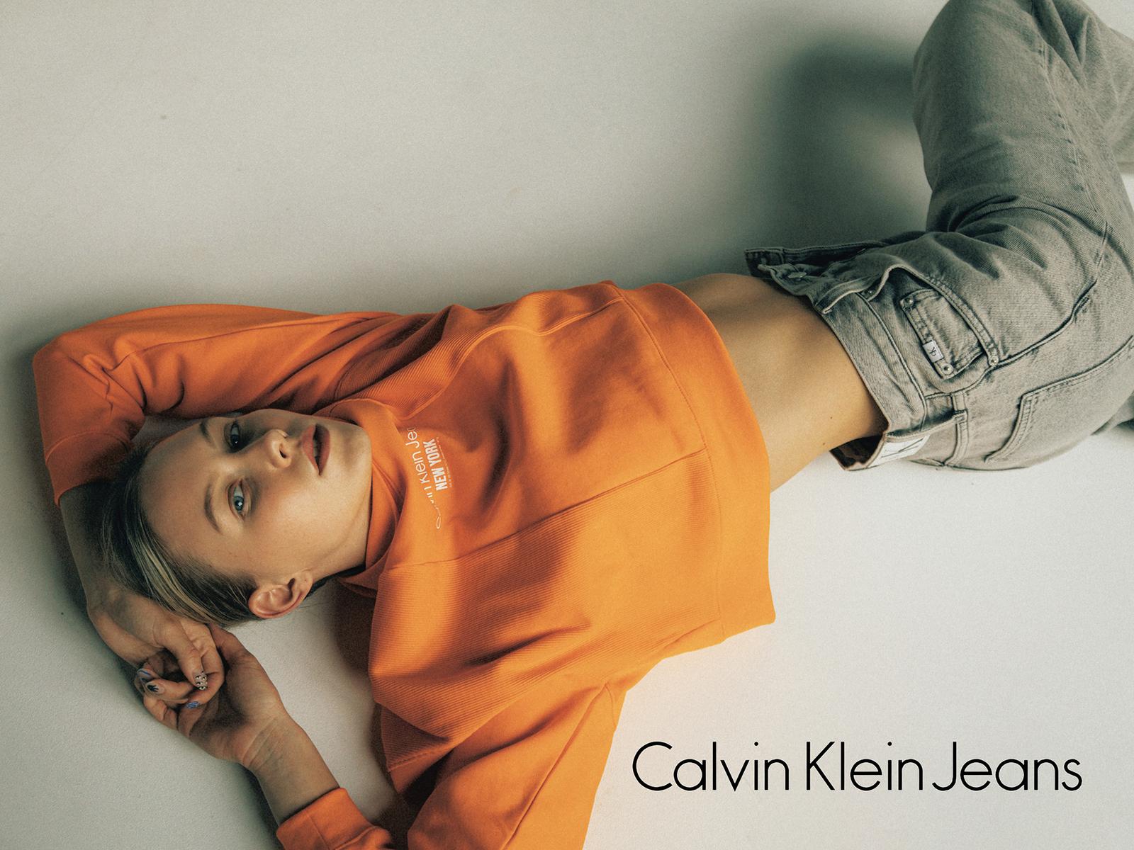 A woman in a bright orange Calvin Klein sweatshirt and jeans lies on the floor, looking directly at the camera with a relaxed expression.