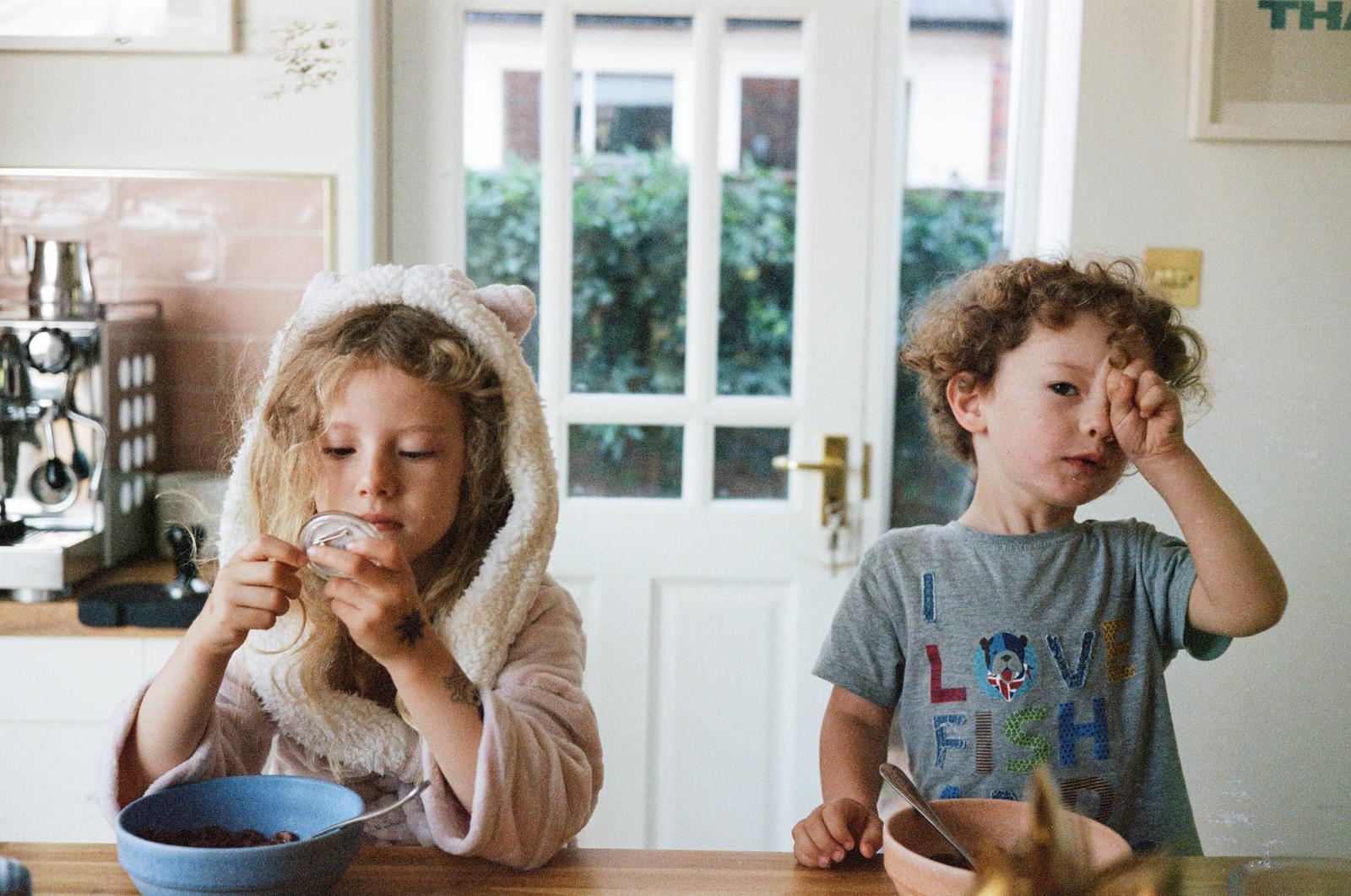 Two children sit at a table, eating cereal. The girl is wearing a fuzzy white robe with a hood.