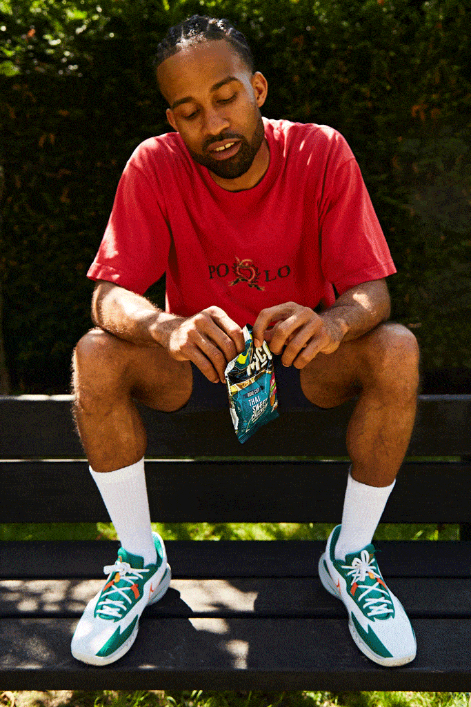 A man sitting on a bench, wearing a red polo shirt, blue shorts, and white sneakers, holding a bag of chips.