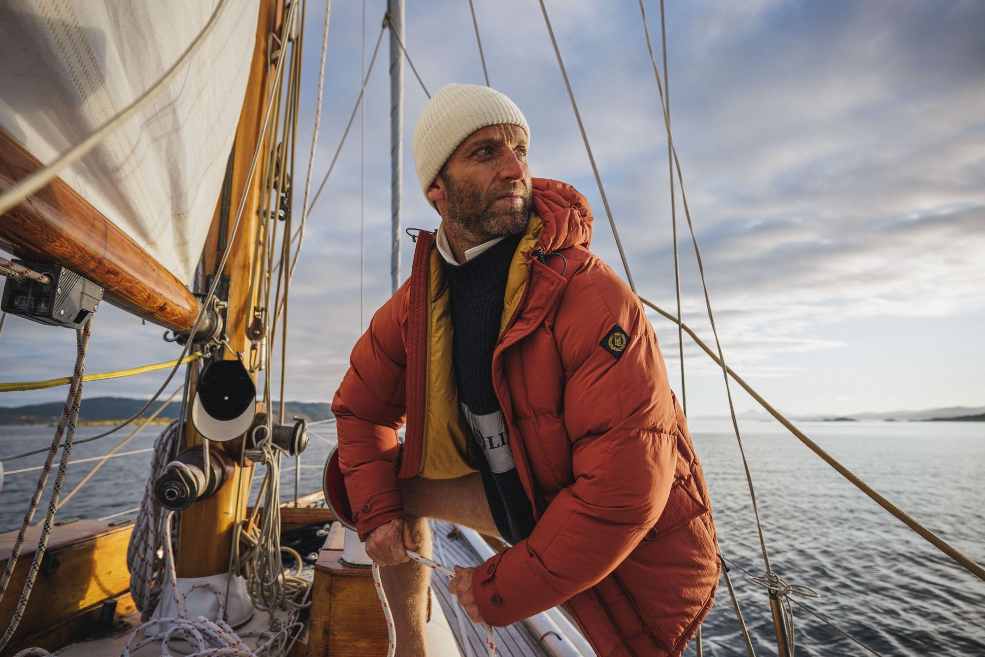 A man in a red jacket and white beanie standing on a sailboat, looking out at the sea.