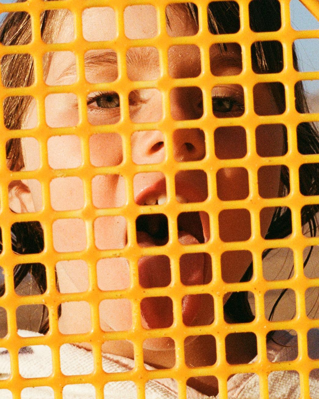 A close-up of a woman's face through a yellow plastic grid, with her eyes and hair visible through the holes.