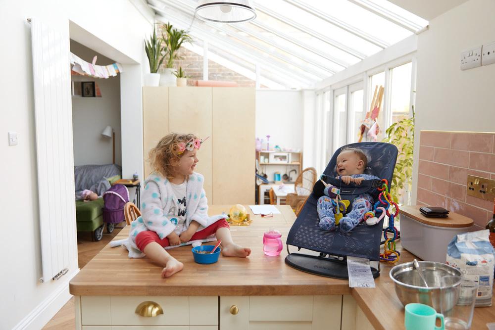 A young girl and an infant sit on a kitchen counter, laughing and playing together. The girl, wearing a white robe with blue polka dots, has a bowl of cereal and a straw in front.
