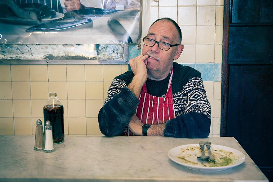 A man in a red and white striped apron sits at a table in a restaurant, looking dejected and defeated.