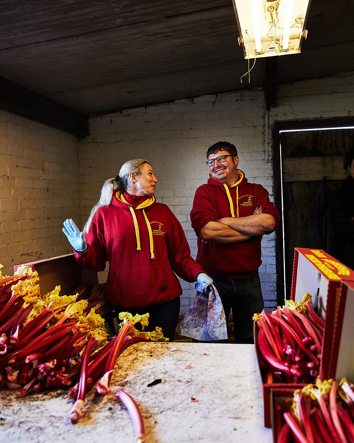 A woman and a man in red hoodies standing near a table with rhubarb in a warehouse.