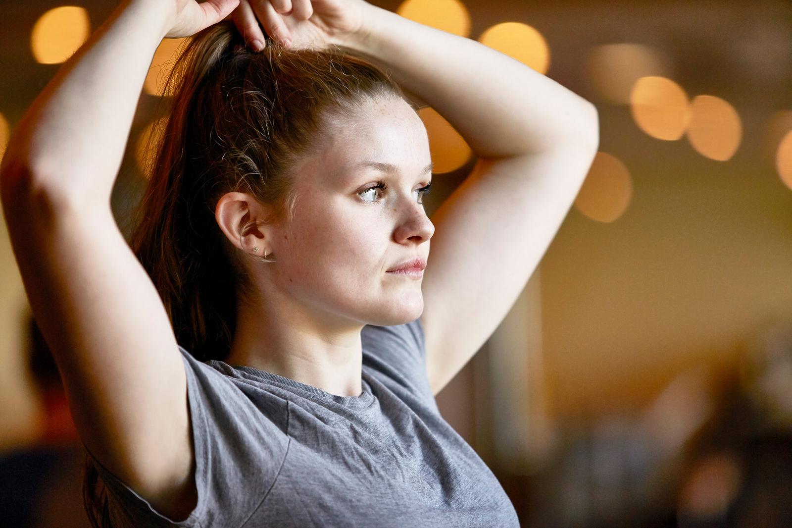A woman with long brown hair, wearing a grey t-shirt, is tying her hair into a ponytail.