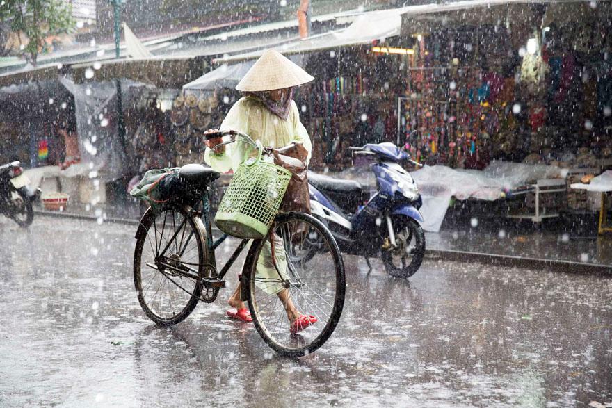 A woman in a traditional Vietnamese conical hat and raincoat is walking in the rain, carrying a bicycle and a basket.