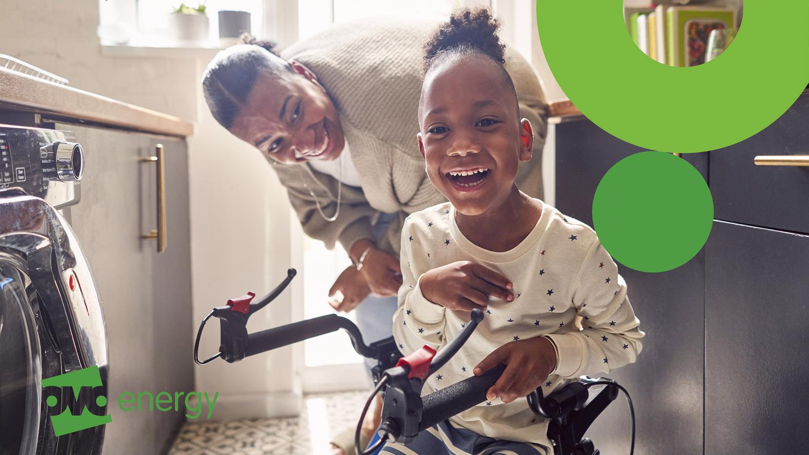 A woman and a young boy in a kitchen, with the boy on a child's bicycle, both smiling and looking at the camera.