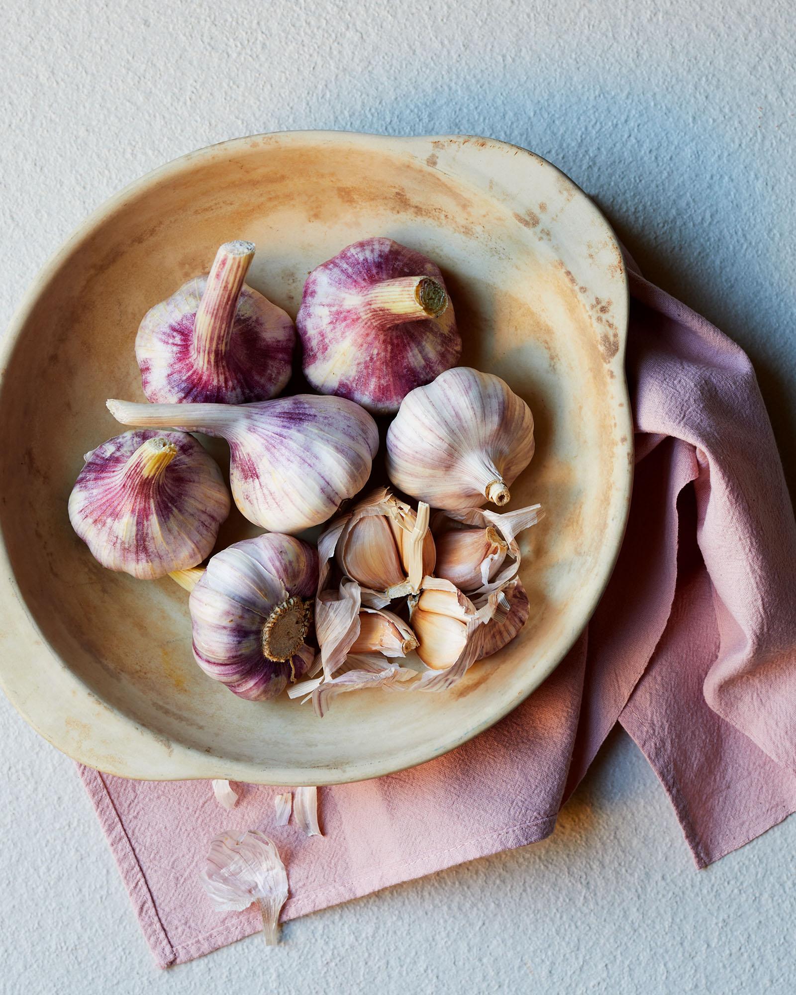 A bowl of variously-sized, multi-colored, and multi-patterned garlic bulbs, some with their tops removed, others still whole, all arranged in a shallow, off-white, ceramic bowl.