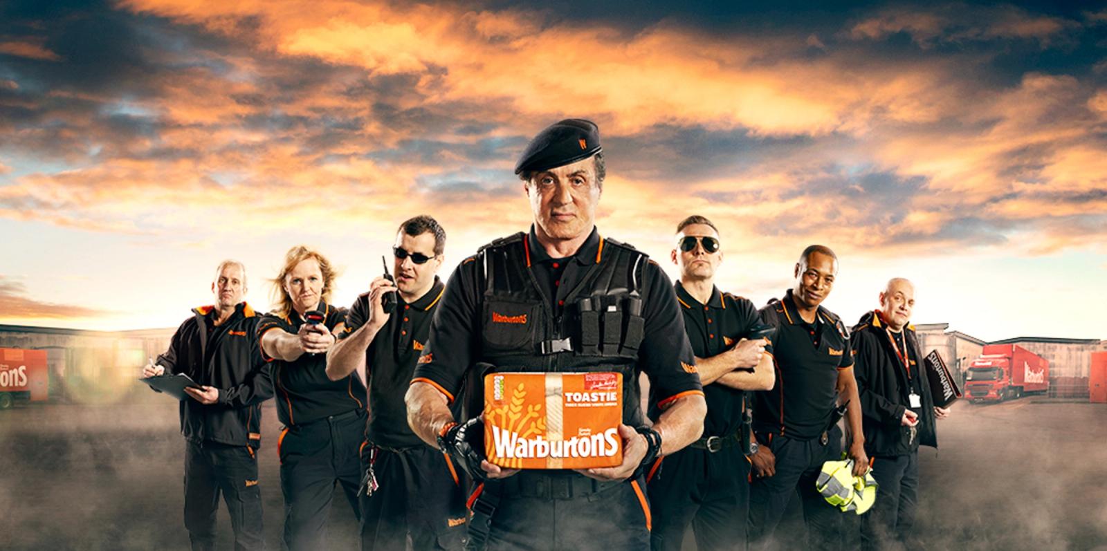 A group of people in black and orange uniforms, holding various items, including a Warburtons box, in a warehouse setting.
