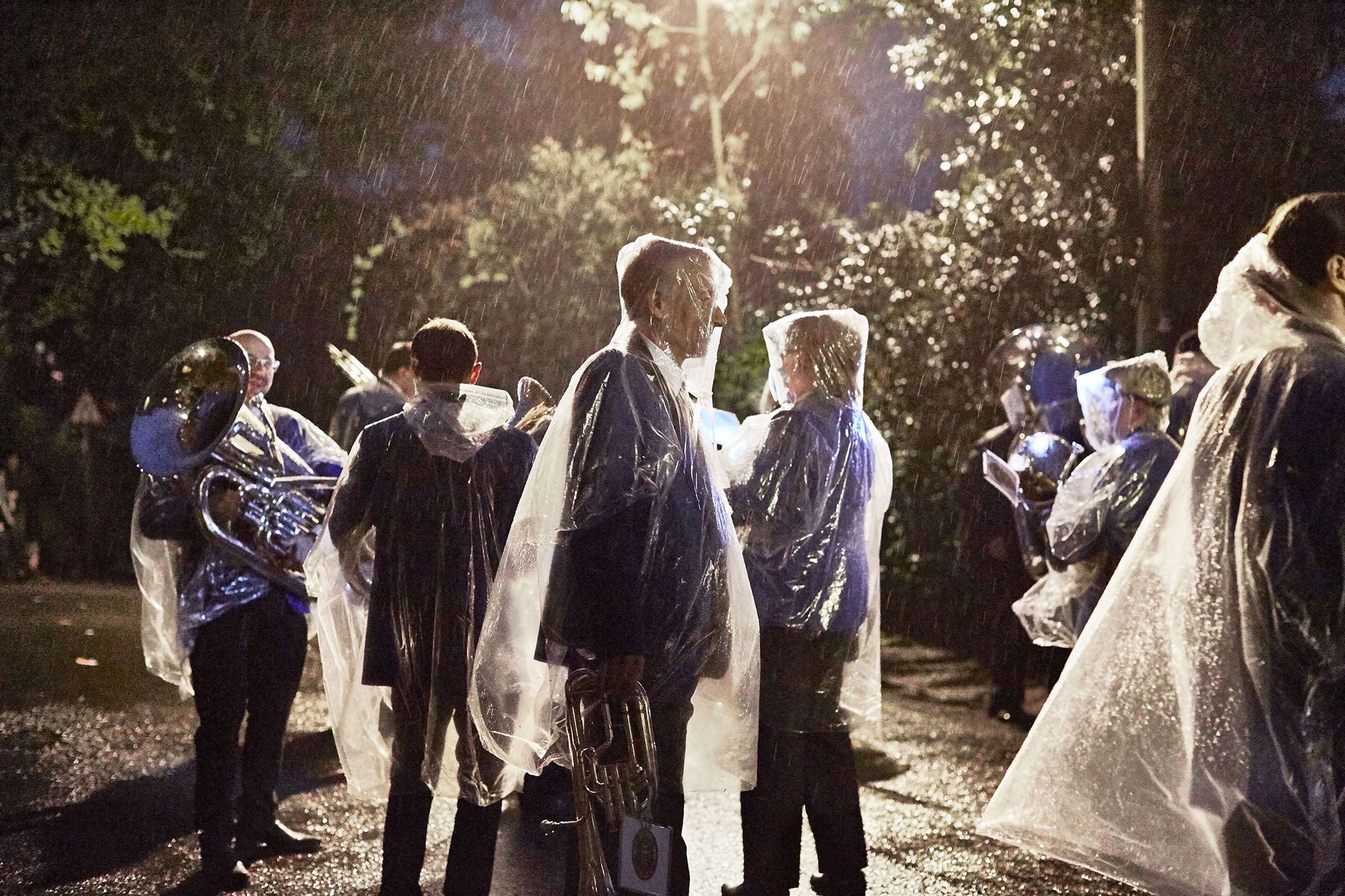 A group of people, likely a band or orchestra, standing in the rain, wearing raincoats and holding musical instruments, in a dark and rainy outdoor setting.
