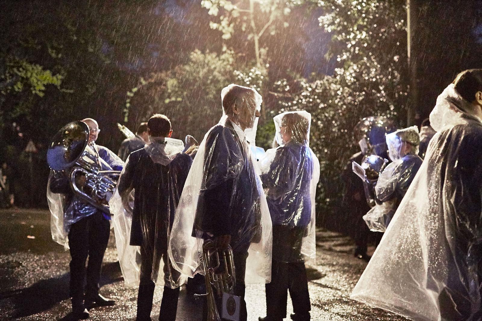 A group of people, likely a band or orchestra, standing in the rain, wearing raincoats and holding musical instruments, in a dark and rainy outdoor setting.