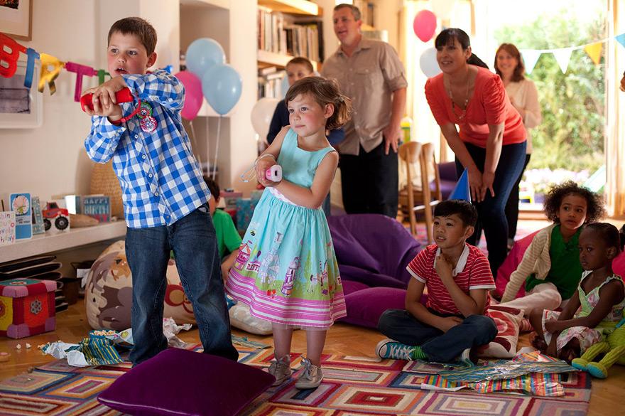Children and adults in a room with toys and decorations, some standing on a purple cushion while others sit on bean bags, all looking at something off-frame.