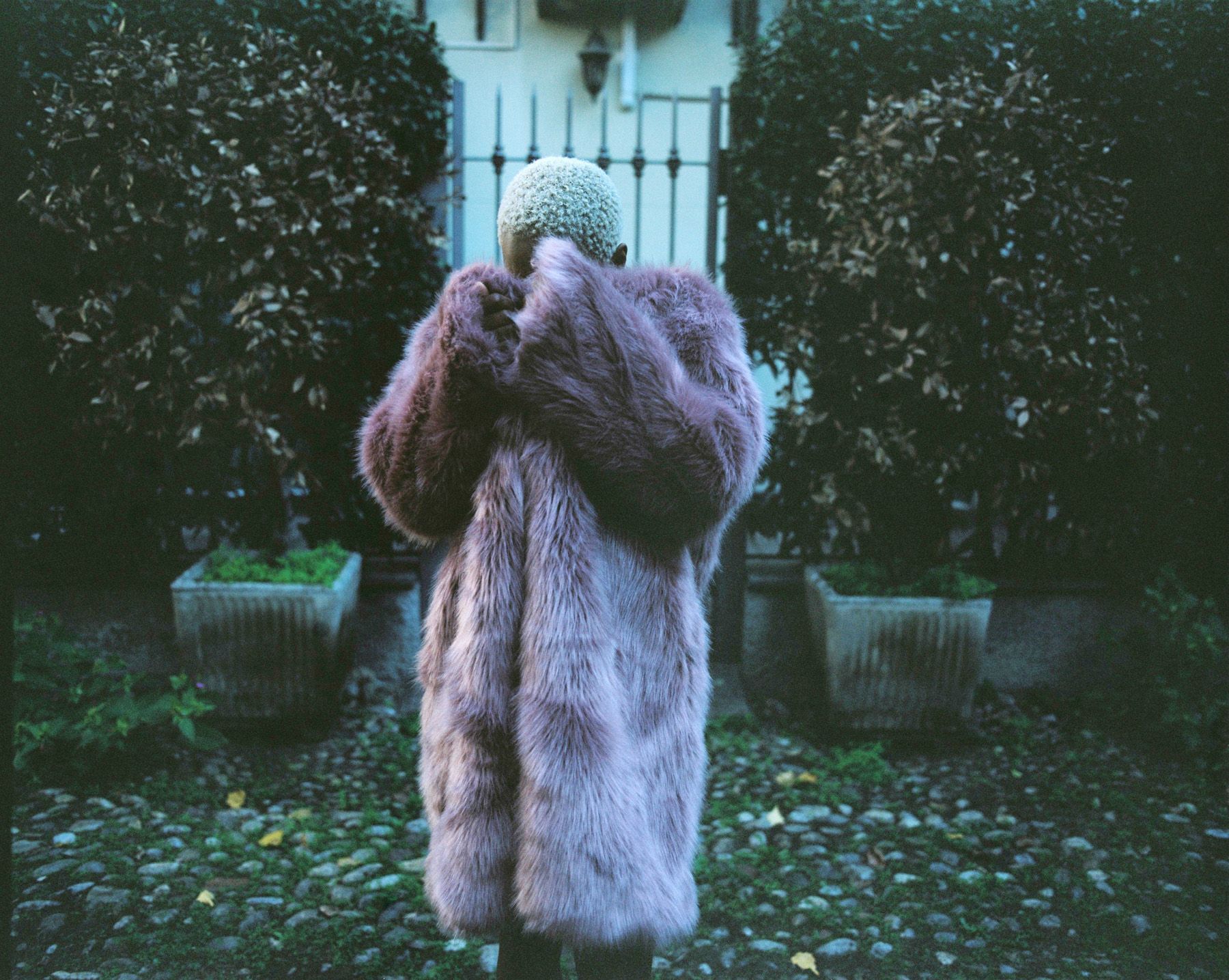 A person wearing a long, fluffy, pink fur coat, standing in a garden with a stone path and a metal fence in the background.