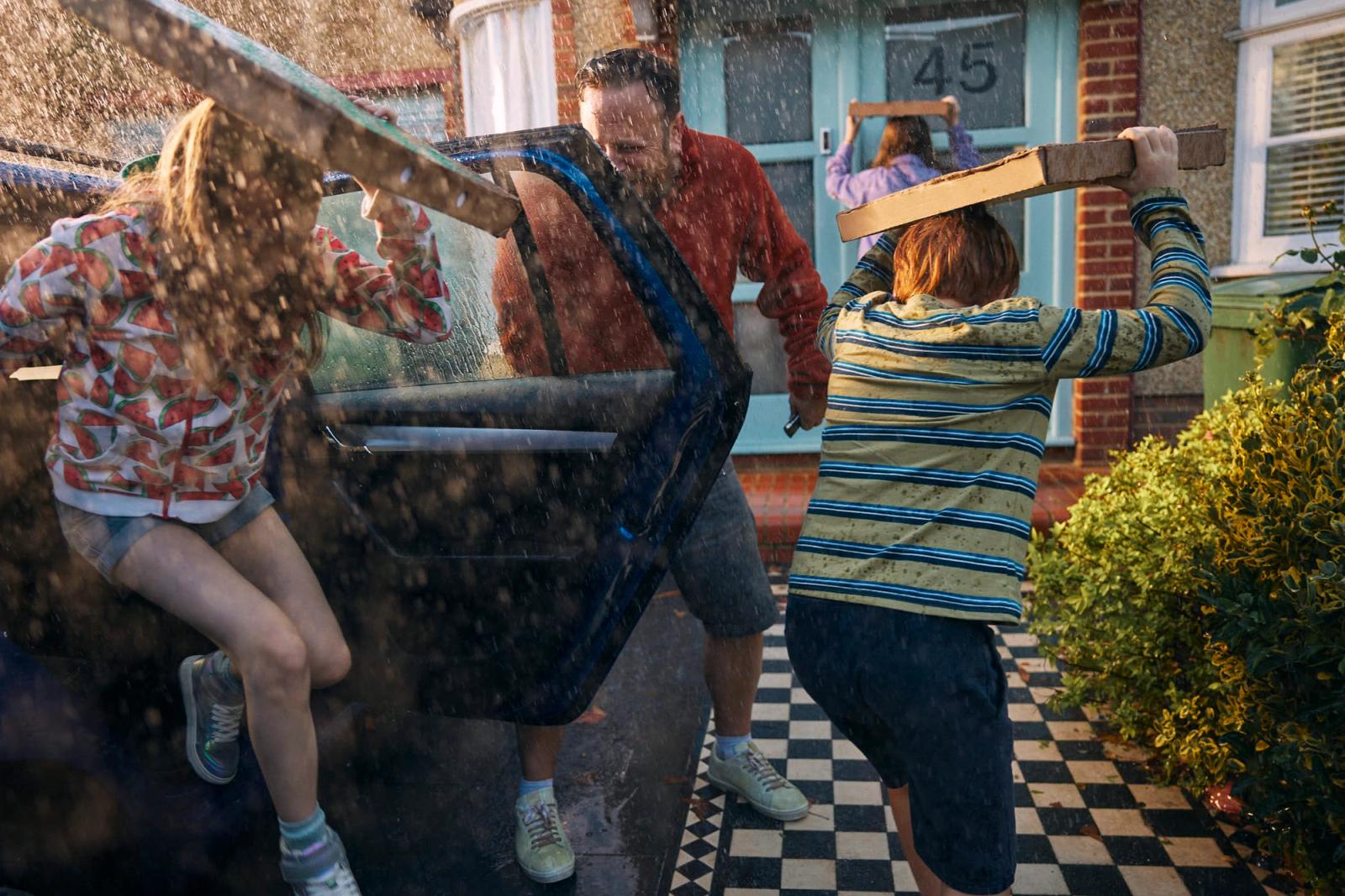 A family of four, including two children, is caught in a sudden downpour as they try to load a car. The father, wearing a red shirt and shorts, is struggling to close the car.