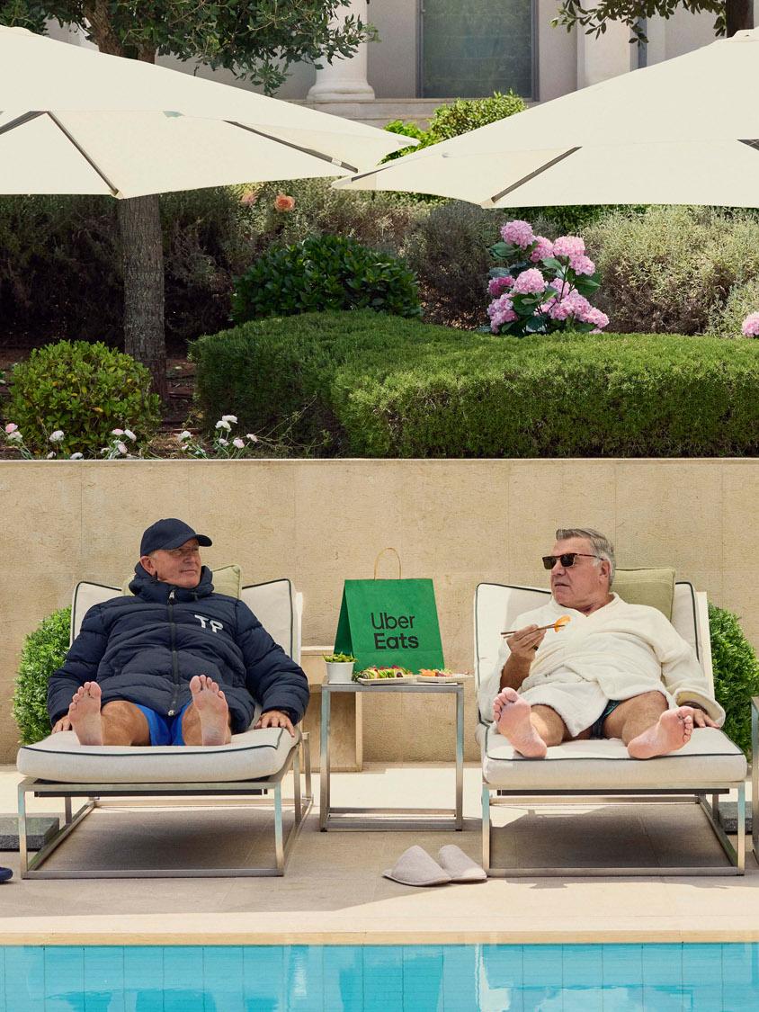 Three older men in bathrobes and slippers lounging by a pool, surrounded by food and Uber Eats delivery bags.