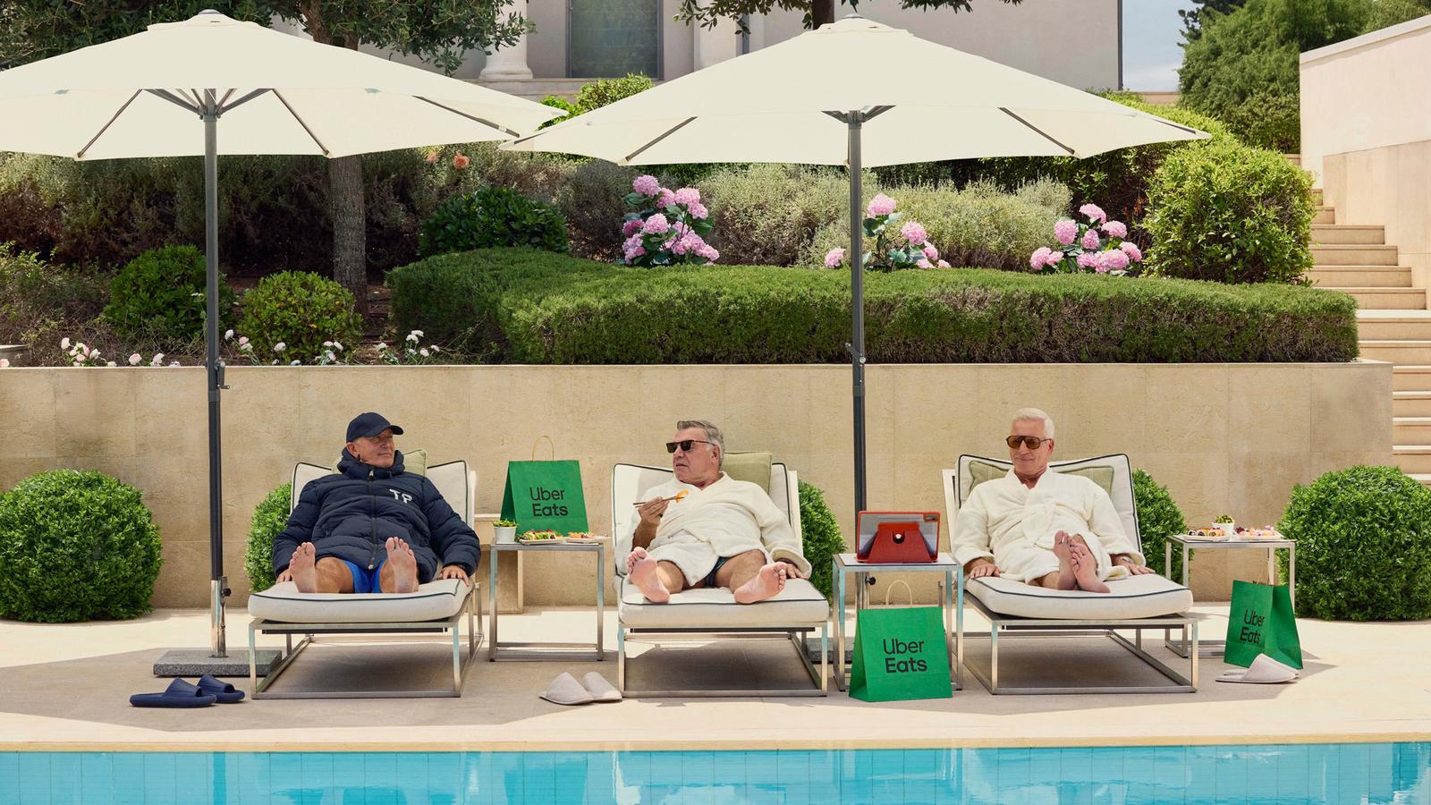 Three older men in bathrobes and slippers lounging by a pool, surrounded by food and Uber Eats delivery bags.