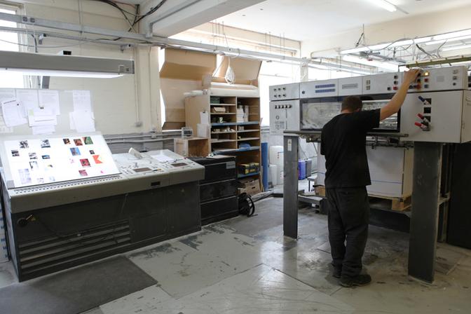 A person in a black t-shirt and jeans standing in front of a large printing machine, with various screens and controls in the background.