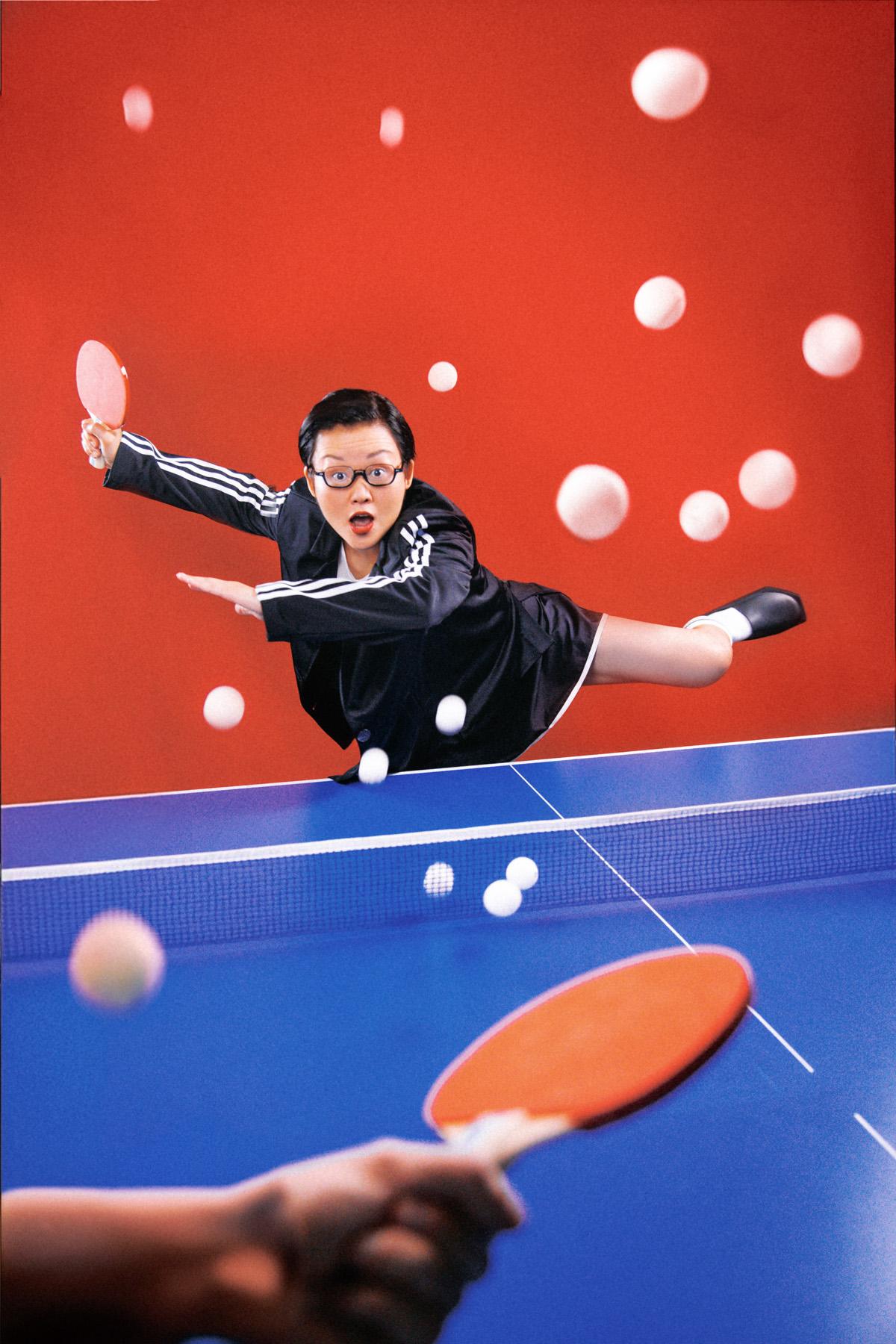 A woman in a black and white Adidas tracksuit is in mid-air, jumping over a table tennis table, with a table tennis paddle in her hand.