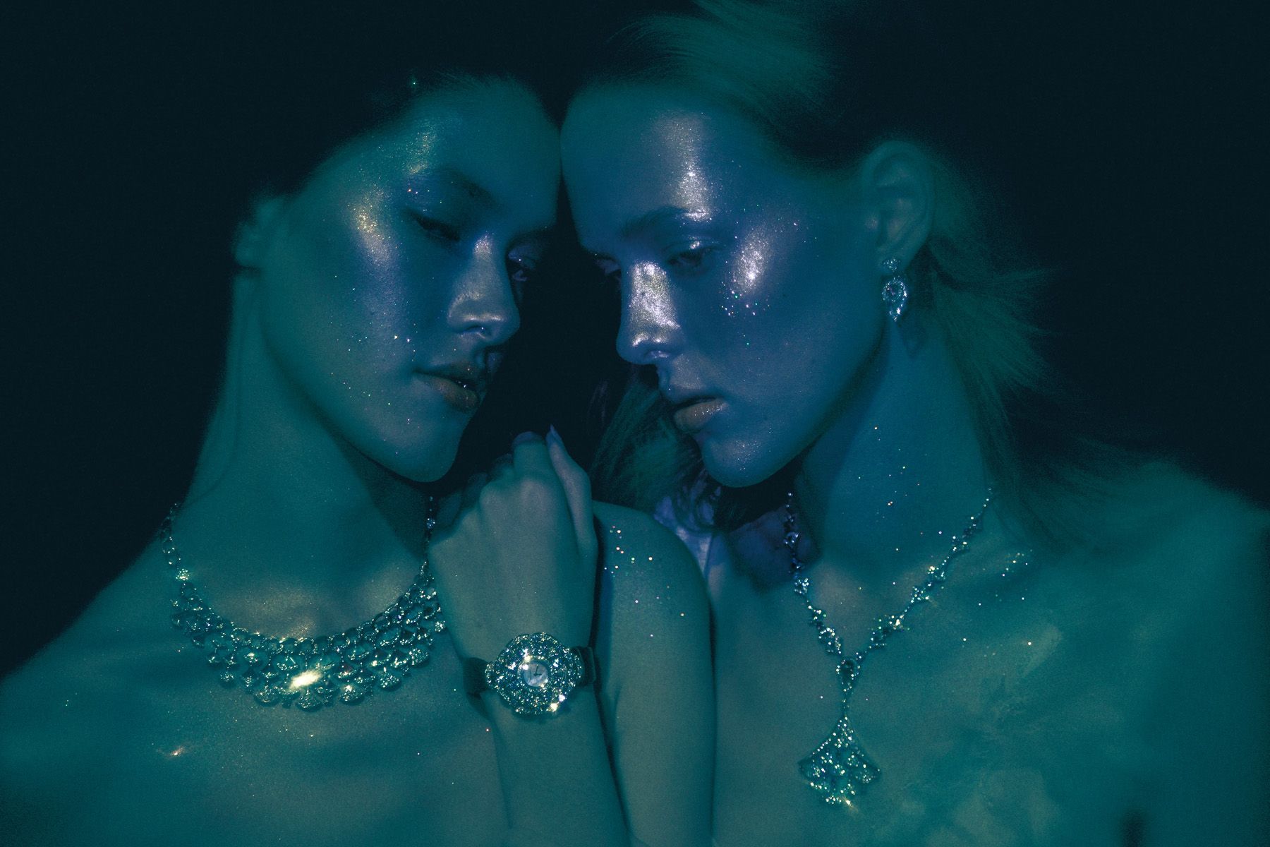 Two women embrace, touching foreheads, in a dark, moody setting, faces and bodies covered in shimmering substance.