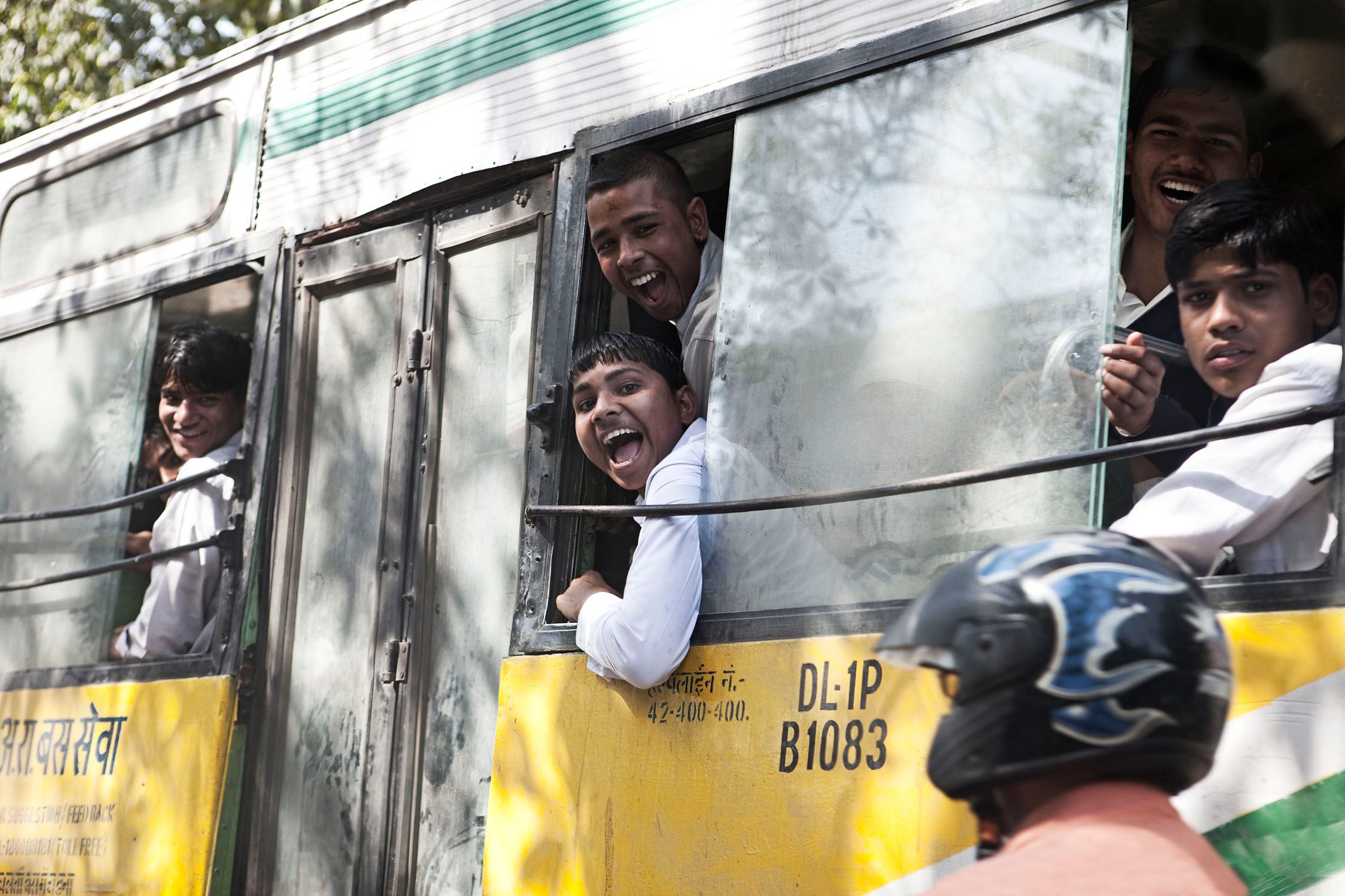 A group of young men in white shirts are looking out of a bus window, with one of them sticking his head out of the window and making a face.