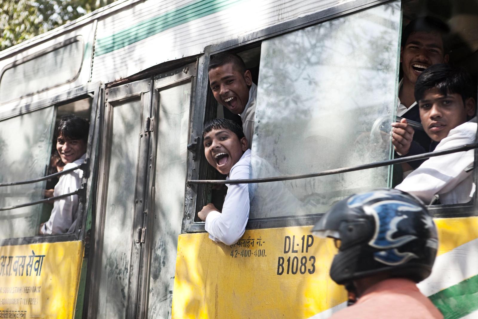 A group of young men in white shirts are looking out of a bus window, with one of them sticking his head out of the window and making a face.