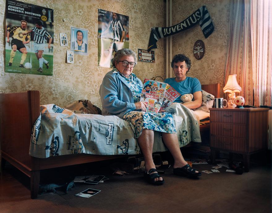 An older woman and a man are sitting on a bed, surrounded by football posters and other football-related items, with the woman holding a magazine and the man holding a teddy bear.
