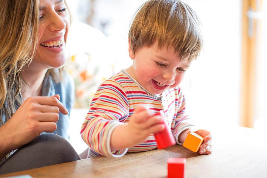 A young woman with long blonde hair is sitting next to a toddler boy who is playing with red and orange building blocks on a wooden table.