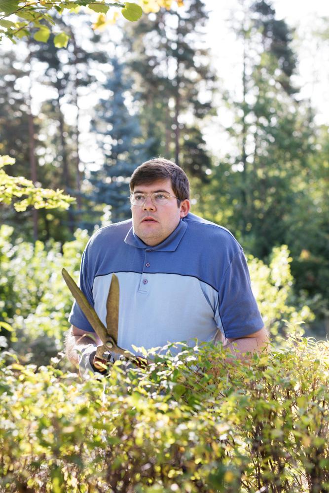 A man in a blue and white striped polo shirt, holding a pair of large, rusty garden shears, standing in a garden or forest, surrounded by greenery.
