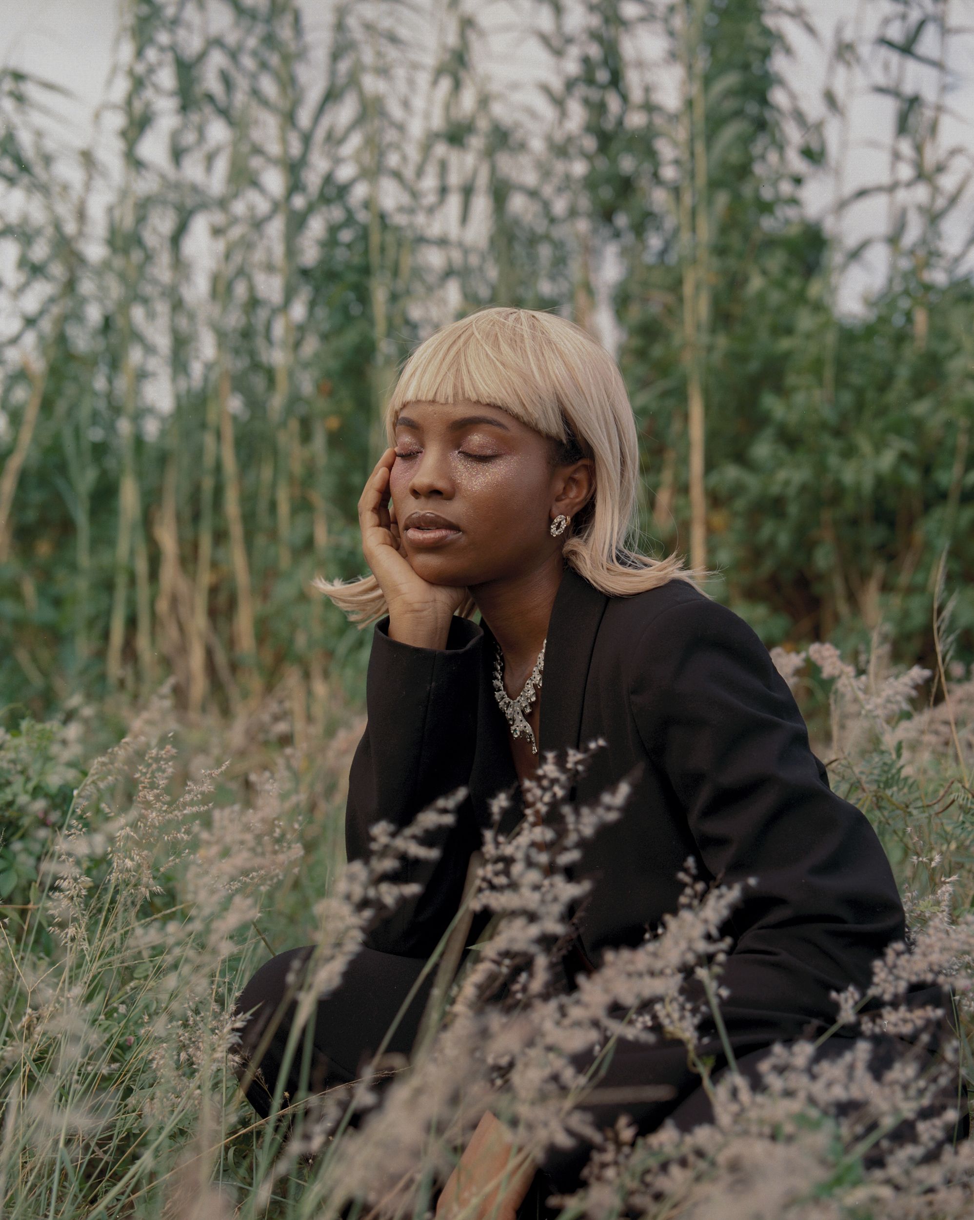 A woman with blonde hair and a black blazer sitting in a field of tall grass and wildflowers, wearing a black blazer and a silver necklace.