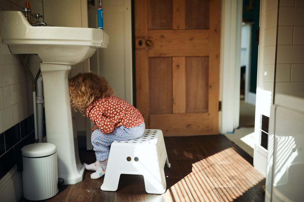A young girl is sitting on a white stool, peering into a bathroom sink. She is wearing a red and white floral top, blue and white floral pants, and has curly blonde hair.