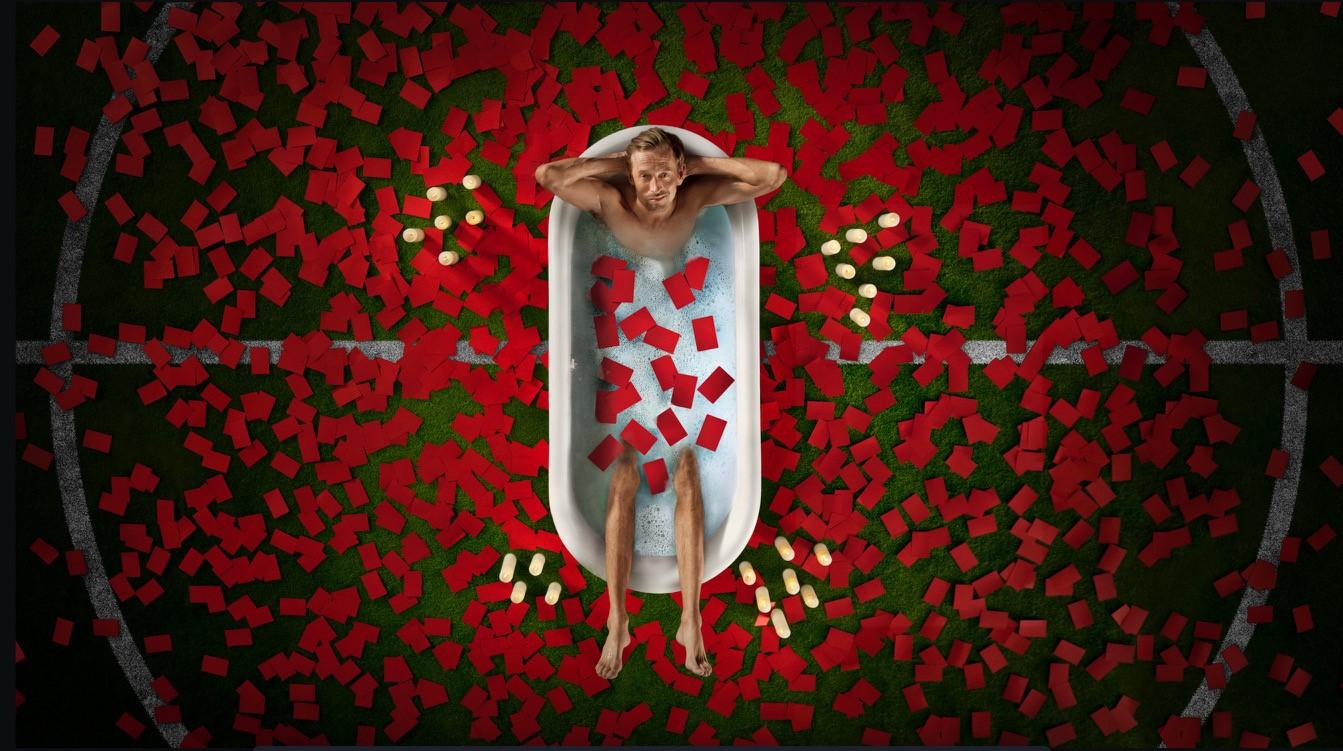 A man lies in a bathtub, surrounded by red rose petals and white flowers, with his eyes closed and a peaceful expression on his face. He is completely submerged in the water,.