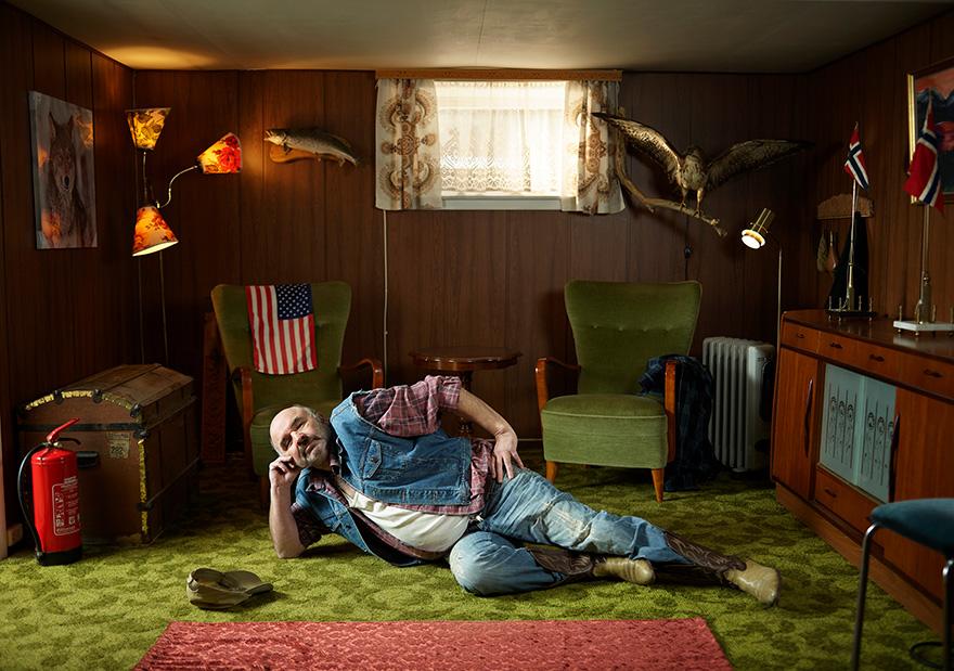 A man lies on a green carpeted floor surrounded by objects including a fire extinguisher, TV, and chair.