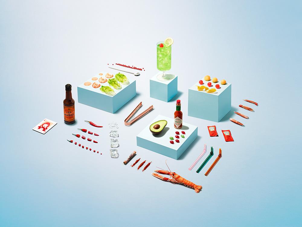 A still life image featuring a glass of green liquid, a bottle of hot sauce, an avocado, and a lobster.
