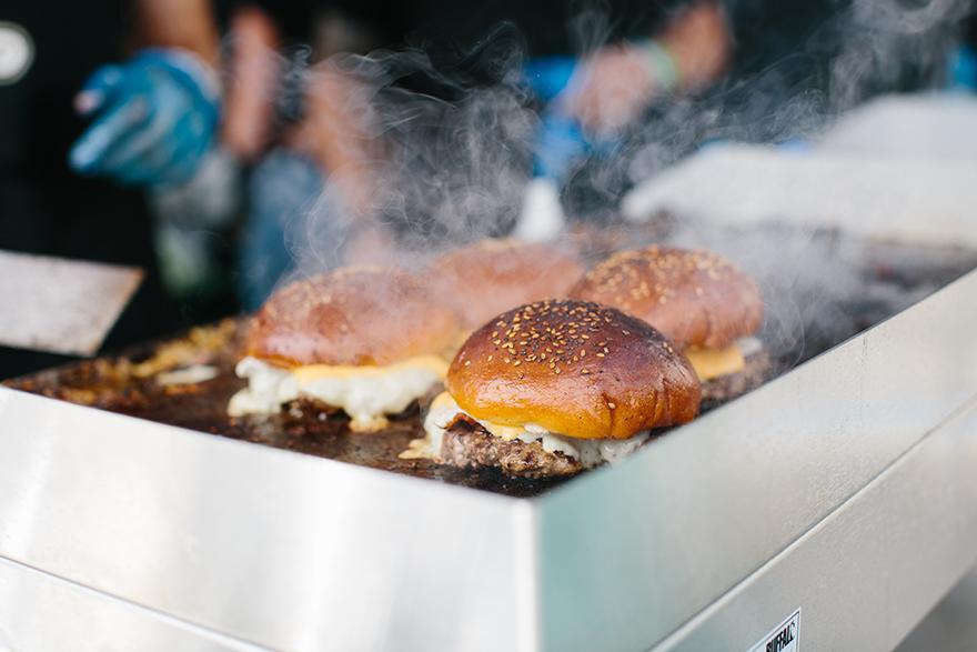 A large, industrial-style grill is filled with multiple, large, beef burgers, each topped with melted cheese and a variety of condiments, being cooked to a golden brown.