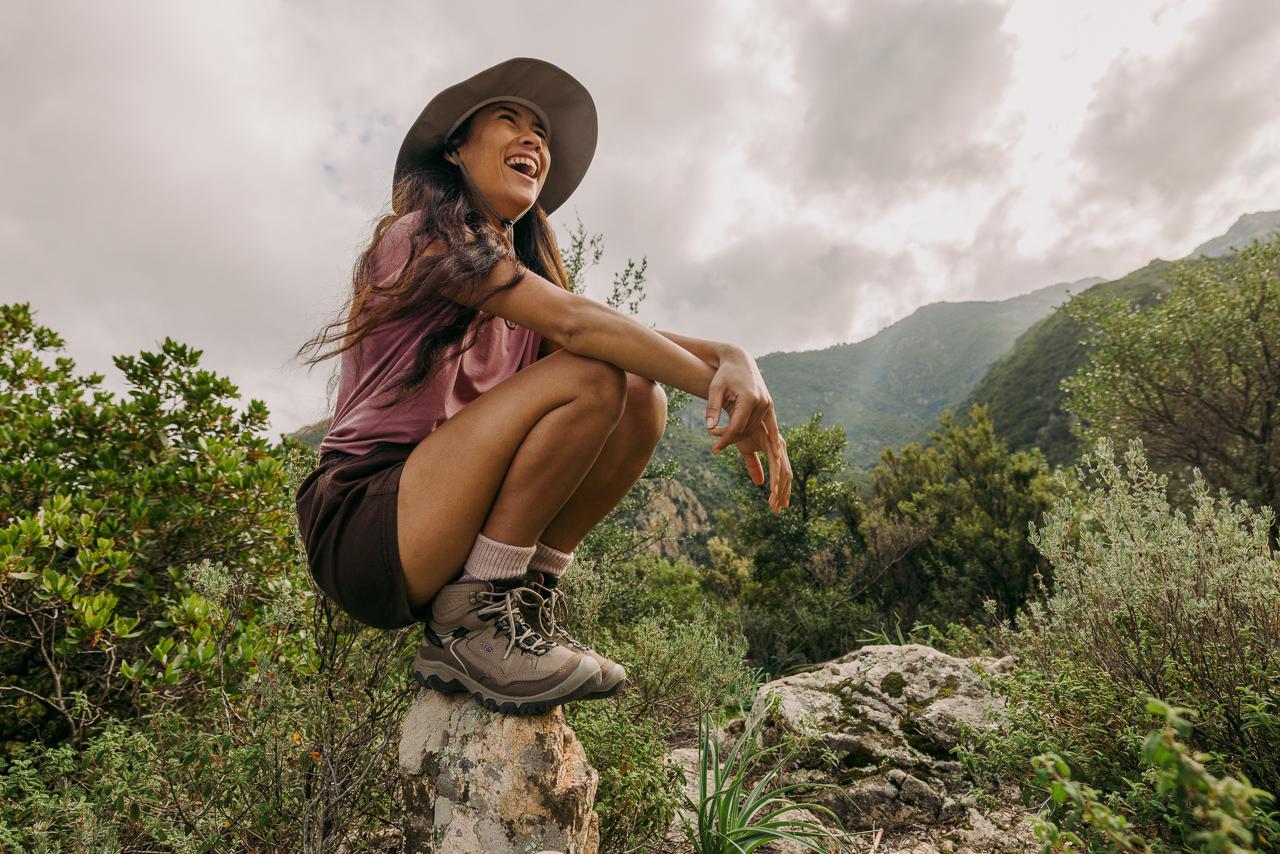 A woman in a wide-brimmed hat, hiking boots, and a pink t-shirt is sitting on a rock in a mountainous area, surrounded by trees and bushes.