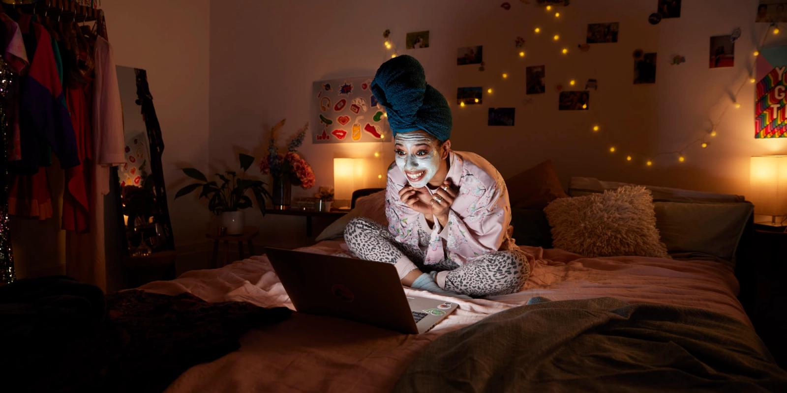 A woman lies in bed, wearing a face mask and pajamas, with her laptop on her lap. She is smiling and looking at the screen, surrounded by a cozy and darkly lit bedroom.