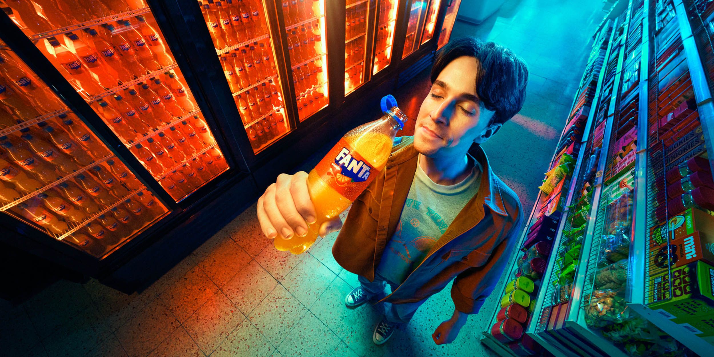 A man stands in front of a wall of refrigerators, holding an Fanta bottle in his right hand. He is wearing a white t-shirt under a brown buttoned-up jacket, and has dark hair. He. gary salter