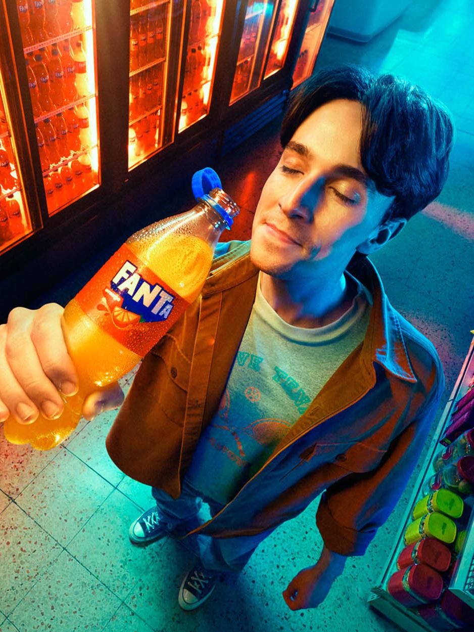 A man stands in front of a wall of refrigerators, holding an Fanta bottle in his right hand. He is wearing a white t-shirt under a brown buttoned-up jacket, and has dark hair. He. gary salter