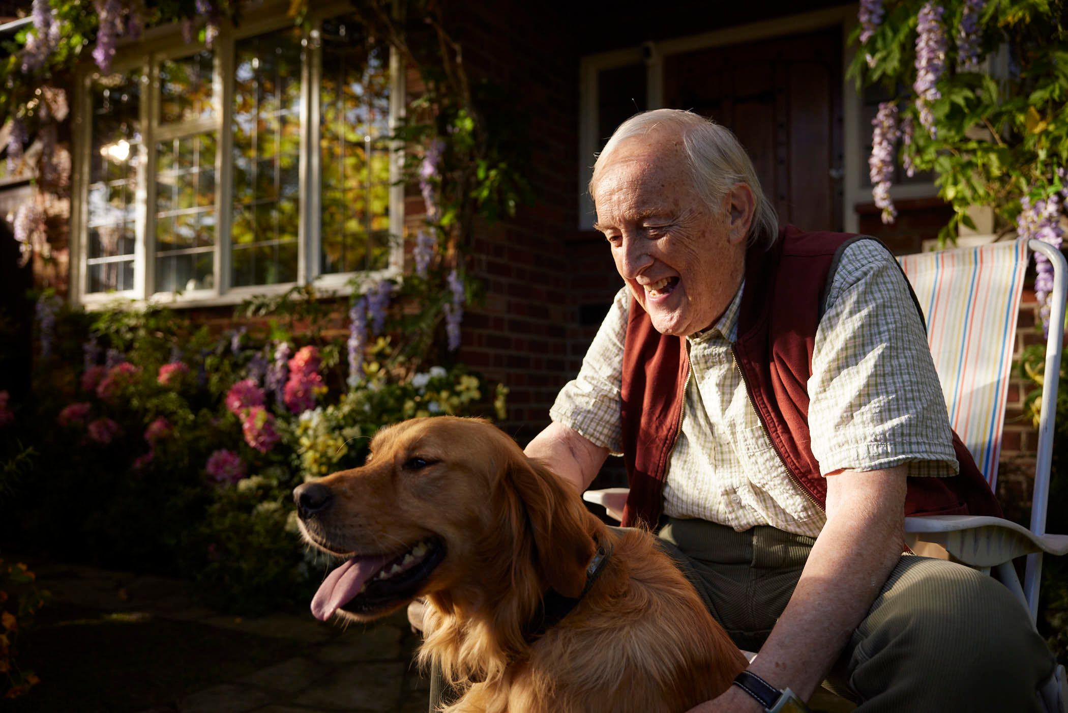 An elderly man sits in a chair with a golden retriever on his lap, both smiling and looking to the left. They are in front of a brick house with a garden full of flowers.
