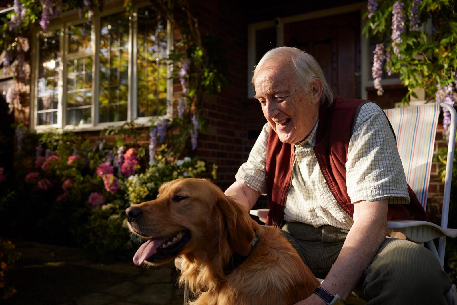 An elderly man sits in a chair with a golden retriever on his lap, both smiling and looking to the left. They are in front of a brick house with a garden full of flowers.